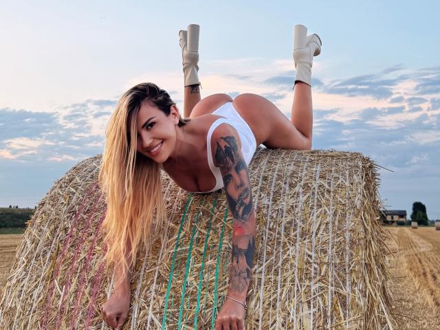 ZoeyMarie with long blonde hair, white tank top, and white panties, lying on hay bale, smiling, showing her big ass and tattooed arm, wearing white boots.
