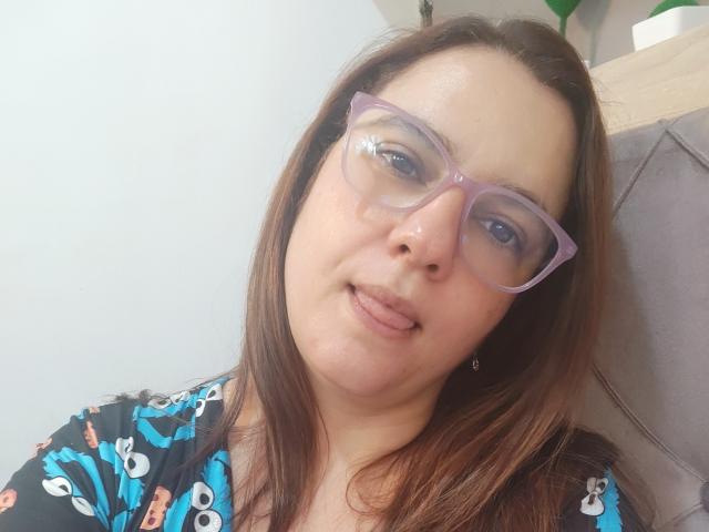Amelia Wilson ha i capelli castano medio, occhiali viola e un viso leggermente lucido con un sorriso appena accennato, la lingua leggermente fuori, indossa una maglietta nera con motivi a cartoni animati blu e bianchi..