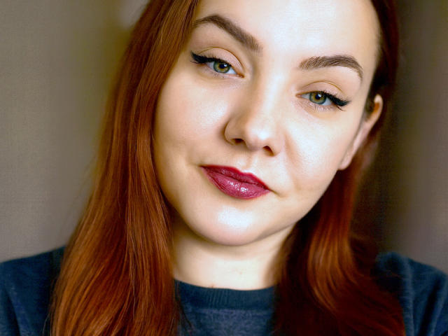 VirgieEuphie has pale skin, red hair, and green eyes with black eyeliner. She's wearing pink lipstick and a dark sweater, smiling slightly with a cute, sexy face.