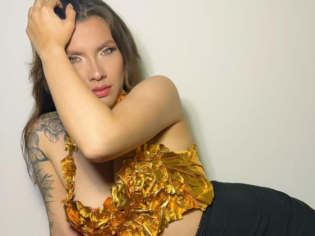 KarlaMorriss with long brown hair, green eyes, and a tattoo on her right arm, wearing a shiny gold top, black pants, and resting her hand on her head.