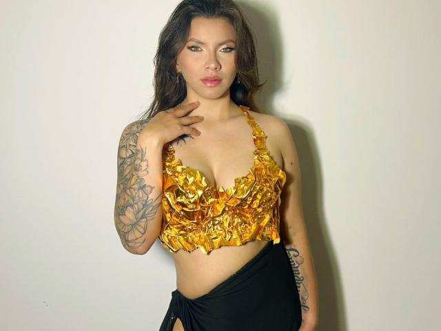 KarlaMorriss with brown hair, yellow ruffled crop top, black skirt, tattoos on right arm, hand on chest, standing against white wall, looking at camera with a slight smile.
