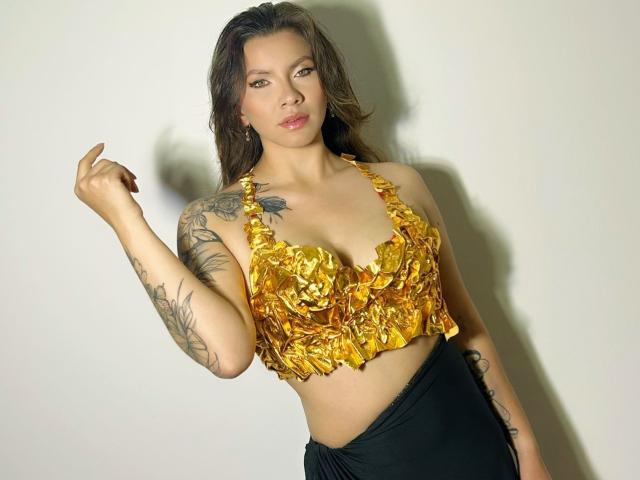 KarlaMorriss with golden, leaf-patterned top, showing tits, black skirt, tattoos on arms, brown hair, neutral expression, holding finger up, white background.