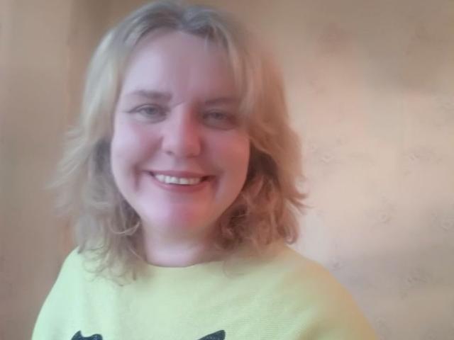 VestaWoody has blonde, wavy hair, a cute smile, and is wearing a yellow shirt. Her tits are small, and she's looking directly at the camera.