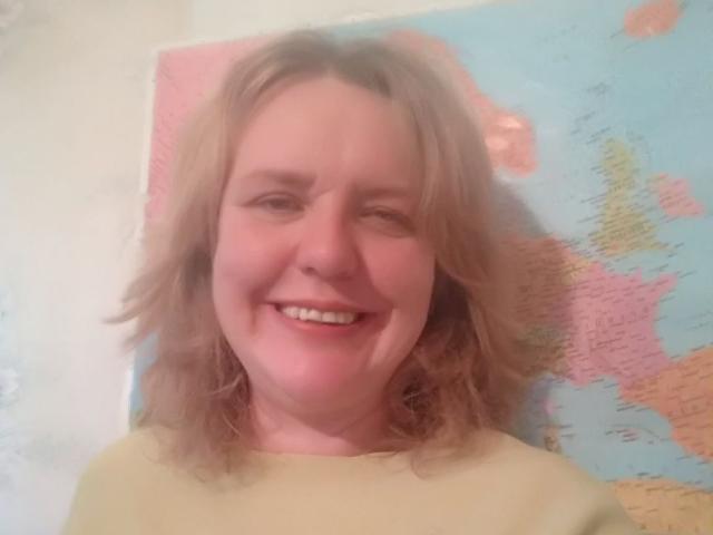 VestaWoody has blonde, wavy hair, a fair skin tone, and a friendly smile, wearing a yellow shirt, with a map behind her. She looks cute and happy.