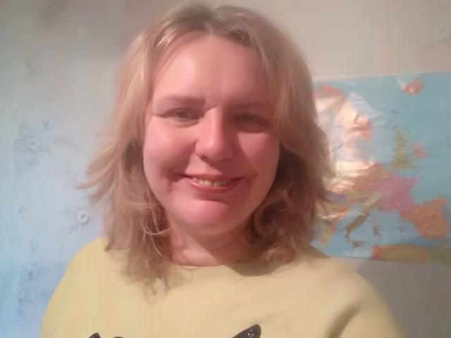 VestaWoody has blonde, slightly wavy hair, a light skin tone, and a small smile. She's wearing a yellow shirt with a black design, and her ass is not visible.