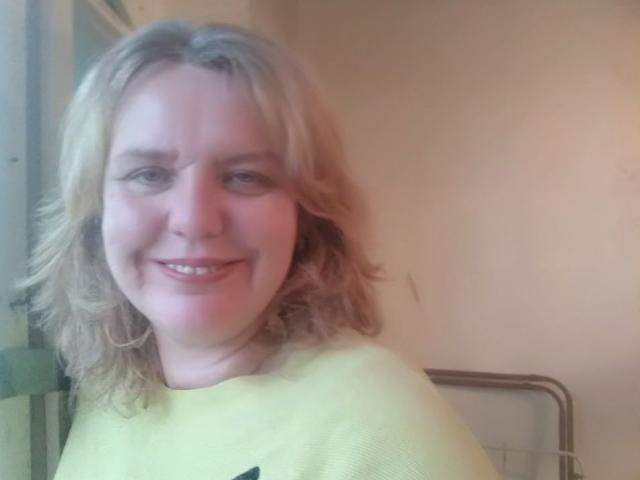 VestaWoody has blonde, shoulder-length hair and a slight smile, wearing a yellow shirt, with light skin and subtle pink cheeks, in a casual, indoor setting.