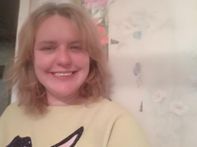 VestaWoody has blonde, wavy hair, a pinkish nose, and is smiling with teeth showing. She's wearing a yellow shirt with a black graphic. Her tits are not visible.
