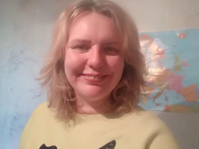 VestaWoody with blonde, wavy hair, smiling, wearing a yellow shirt, has a cute, chubby face, and small tits, standing in front of a map in a slightly blurry photo.