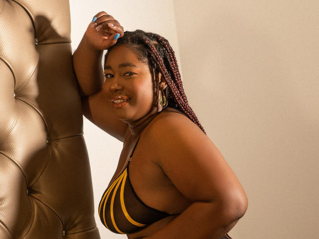 XiomaralLopez, a curvy Black woman with braided hair, leans against a tufted brown wall, wearing a yellow and black striped bikini top, smiling with blue nails.