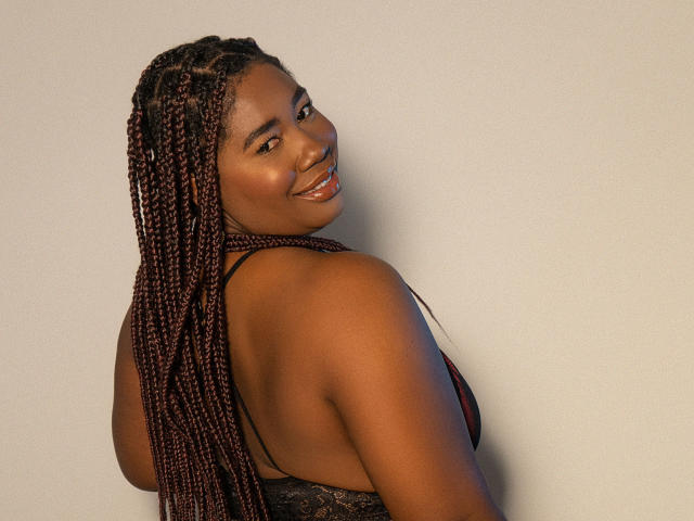 XiomaralLopez with dark brown skin, long braided hair, smiling, looking over shoulder, showing back, wearing black lace-up dress, glowing skin, shiny red lipstick, sexy, confident.
