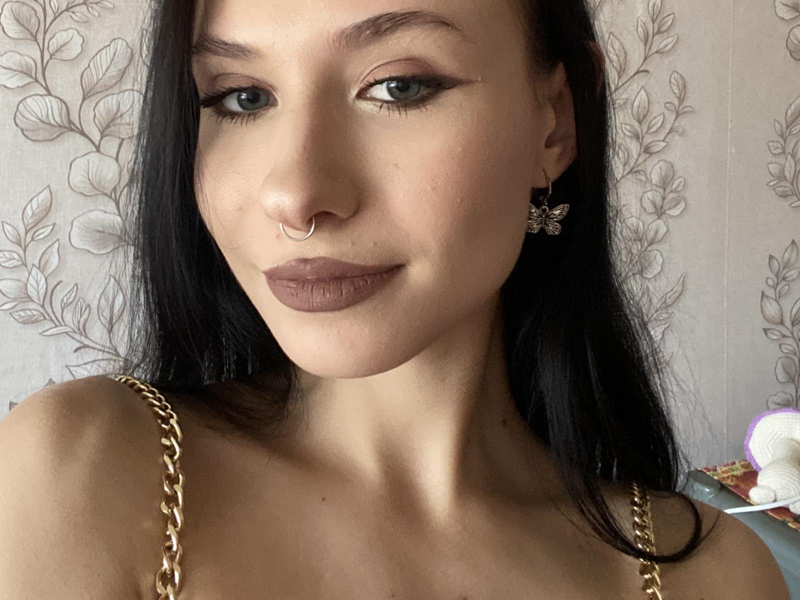 KisaLissa with long black hair, green eyes, and brown lipstick, wearing a gold chain necklace, butterfly earrings, and a nose ring, looking at the camera with a slight smile.