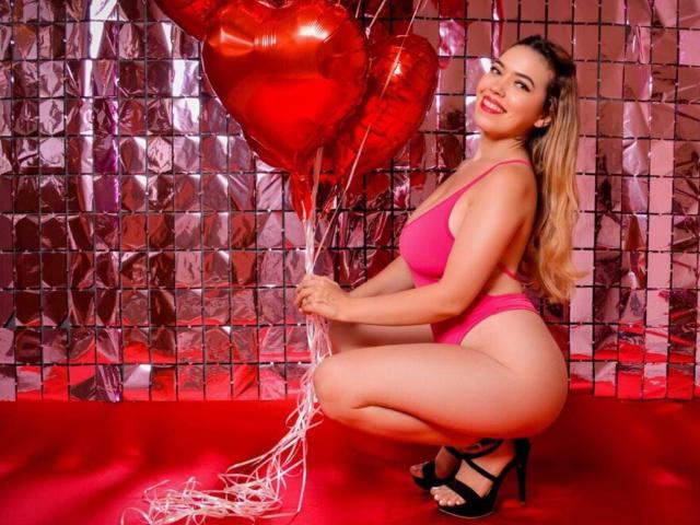 KatieSidney squatting in pink lingerie, holding red heart balloons, smiling, with long blonde hair, black high heels, and a curvy, sexy ass. Red shiny background.