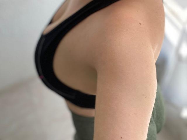 CocoVolkov's arm, showing a tan, smooth, slender upper arm with a black sports bra, green fabric below, and a slight side view of their small, firm tits.