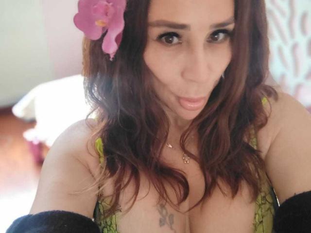 IssaMilfX with brown wavy hair, pink orchid, green lace bra, and black sleeves, pouting with large tits, tattoo below cleavage, looking at camera.