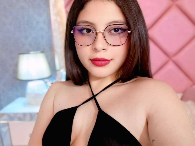 VioletaRoses with glasses, purple eyeliner, red lipstick, black halter top showing cleavage, and medium tits, standing in front of a pink, diamond-patterned wall.