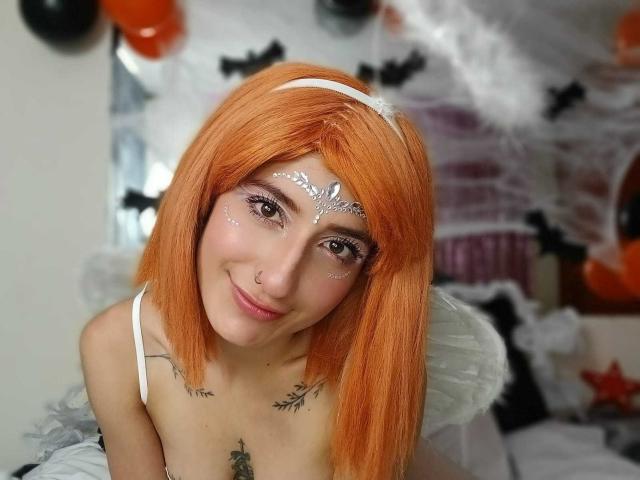 Kaiix con capelli arancioni, fascia bianca per capelli, trucco bianco sul viso, ali bianche, piccolo anello al naso, tatuaggi, sorridente, scollatura visibile, tema di Halloween, illuminazione soffusa, carina, attraente..