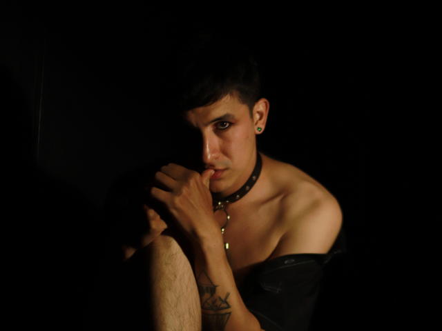 NoahStelle with dark hair, wearing a black choker and off-shoulder shirt, biting his finger, tattoo on right arm, looking serious, dimly lit, shadowy background.