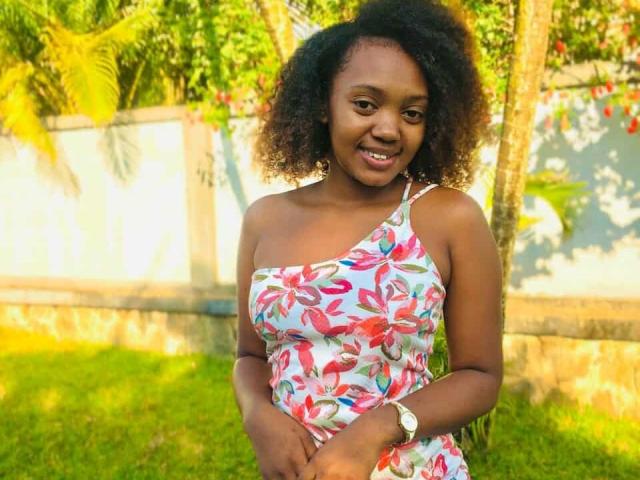 BarbaraSweetz with dark curly hair, smiling, wearing a one-shoulder floral dress, showing her small tits, green grass, and a white watch, standing outdoors in sunlight.