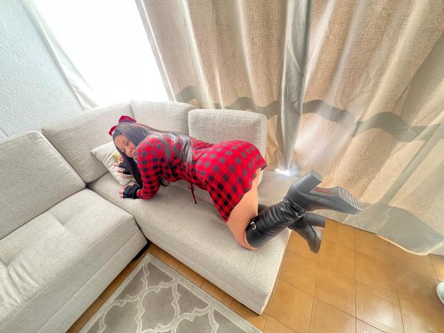 KarinaParker está deitada em um sofá cinza, vestindo um vestido xadrez vermelho, botas pretas e um gorro de enfermeira, mostrando suas nádegas, com a luz do sol entrando pelas cortinas..