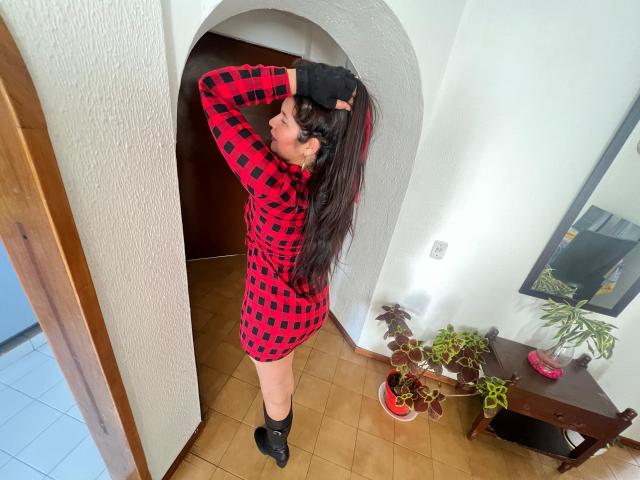 Karina Parker, con lunghi capelli neri, un vestito a scacchi rosso e nero, stivali neri e guanti neri, in piedi nella porta, con le mani tra i capelli, che mostra il sedere, in una stanza con piastrelle..