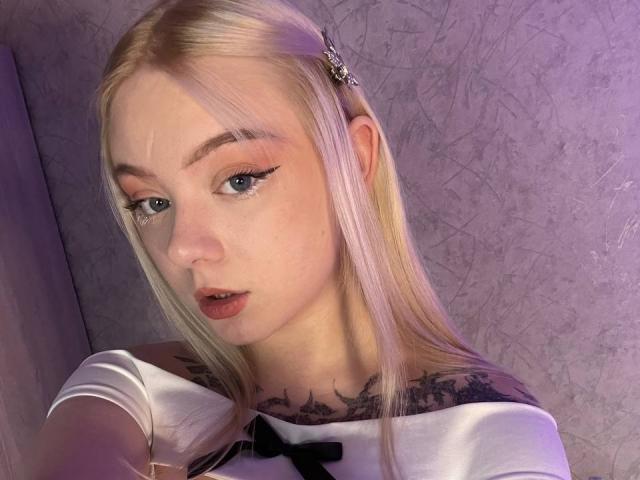 EnchantingEyes with long blonde hair, white off-shoulder top, black bow, eyeliner, tattoos on neck, purple-lit face, and parted lips, looking at camera.