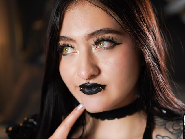 MagicCleo has pale skin, green-yellow eyes with black eyeliner, black lipstick, a nose ring, and a black choker, with long black hair, looking to the side, touching her chin.