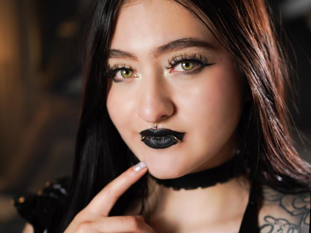 MagicCleo mit grünen Augen, schwarzem Lippenstift, Nasenring und Choker, die mit einem Finger ihr Kinn berührt, hat Tattoos an ihrer Schulter. Ihr dunkles Haar ist glatt und glänzend..