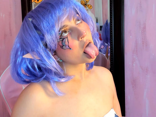 IrinaSweett with blue anime-style hair, pointy elf ears, and a face tattoo, sticking out her tongue, has bare shoulders and a pinkish-white strapless top.