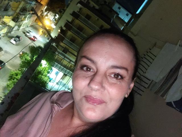 AnaPiyas, a chubby woman with a bald head and dark hair, wearing a pink shirt, has small eyes, glossy pink lips, and a nose piercing. Nighttime city background.