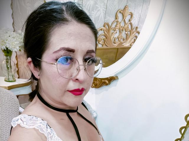 ScarletMilfX mit runder Brille, rotem Lippenstift, schwarzem Choker und weißem Spitzenoberteil, zeigt ein ernstes Gesicht, ihr dunkles Haar ist hochgesteckt. Brüste sind sichtbar, helle Haut, pinke Augen-Make-up..