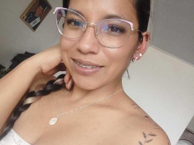 Paulina Colemann, avec des lunettes, des cheveux tressés, souriante, montrant ses dents, portant un collier, de petites boucles d'oreilles, des tatouages sur l'épaule, une main près du visage, peau brun clair, lunettes roses, haut blanc..