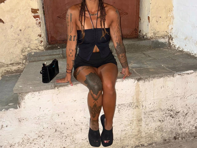 Daphnenights-hot, with tattoos, in a black strapless top and shorts, sitting on a stone ledge, legs crossed, black heels, and black purse, with braided hair and nose ring.
