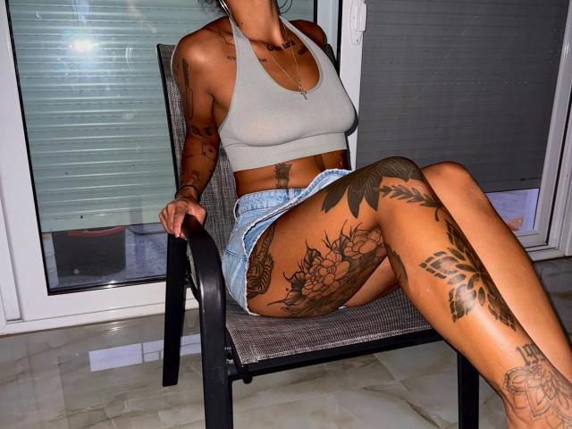 Daphnenights-hot with tattoos, gray sports bra, and denim shorts, sitting on a chair, legs crossed, showing off her tattoos and tan skin, in a brightly lit room.
