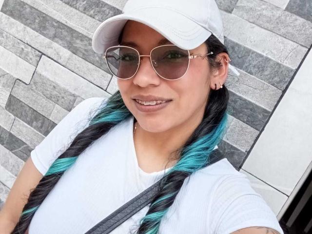 PaulinaColemann has blue and black braids, wearing a white cap, sunglasses, and white tee, with a strap over her tits, smiling with her ass-chop earrings visible.