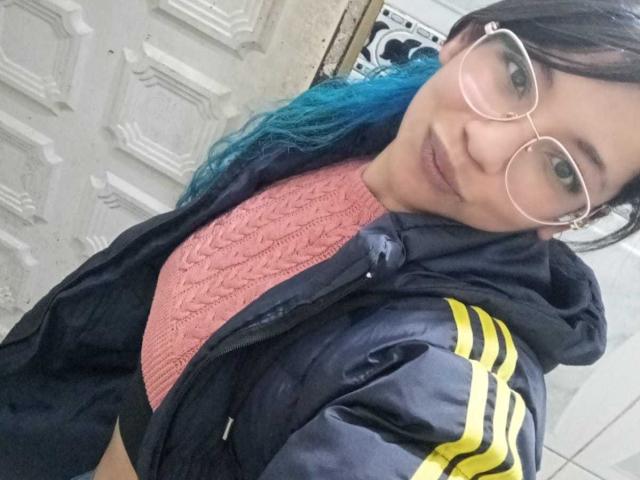 PaulinaColemann with blue-dyed hair, glasses, pink sweater, black jacket with yellow stripes, smiling at the camera, showing her tits under the jacket.