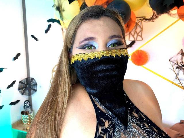SalmaAzaman with green eyes, wearing a black mask with gold lace, and a black, spiderweb-patterned top, showing her bare shoulders and long blonde hair.