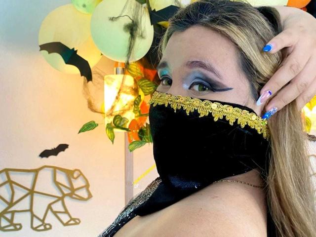SalmaAzaman with dramatic blue eyeshadow, black mask with gold trim, long blonde hair, blue nails, and hand in hair, showing off her tits and a sly smile.