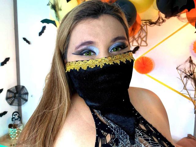 SalmaAzaman with green eyes, blue eyeshadow, black lace mask with gold trim, long blonde hair, black lace top, visible collarbone, and a cute, sexy Halloween vibe.