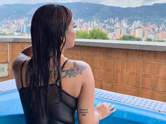 IsabellaMoreau with wet black hair, back to camera, in a blue hot tub, wearing a sheer black top, showing a tattoo on her right shoulder and another on her left arm.
