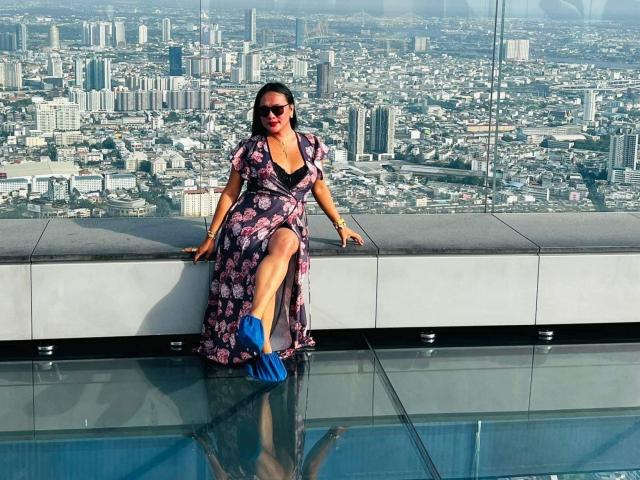 TsPrettyNaughty with dark hair, sunglasses, floral dress, and blue heels, sitting on a glass rooftop, her right leg up, showing a thigh, and her blue heel in water.
