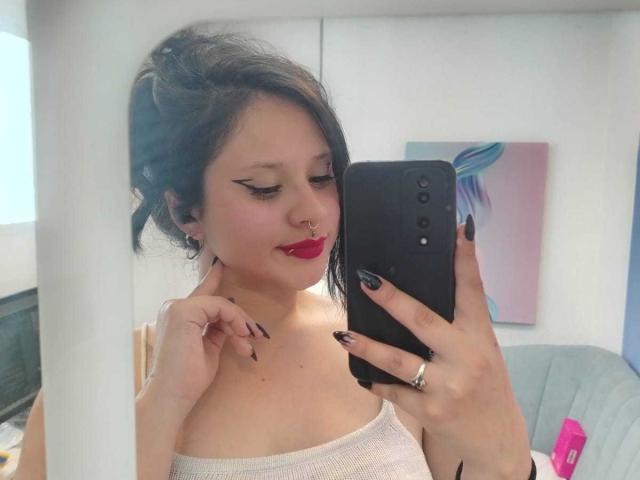 MaraVixen with black nail polish, red lipstick, and nose ring, holding a black phone, taking a mirror selfie, showing her tits in a white strap top.