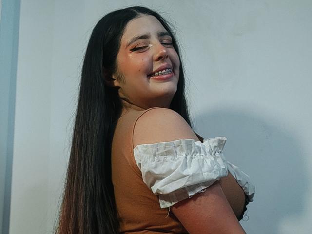 PaolaLyon with long black hair, smiling, wearing a brown off-shoulder top, showing her tits, standing against a white wall, looking over her shoulder.