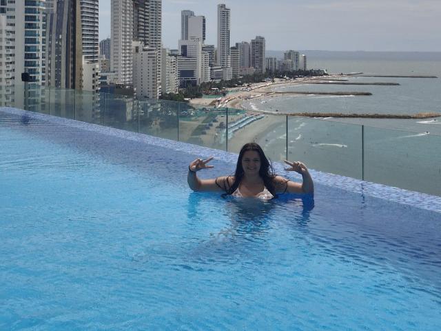KatherinLatin in a blue-tiled infinity pool, water up to her chest, showing off her tits with hands making rock gestures, city skyline and ocean in the background.