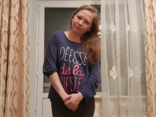 LolaAlina with medium tits, brown hair, wearing a navy blue "Deesse de la vie" shirt, standing in front of a window with sheer curtains, hands crossed.
