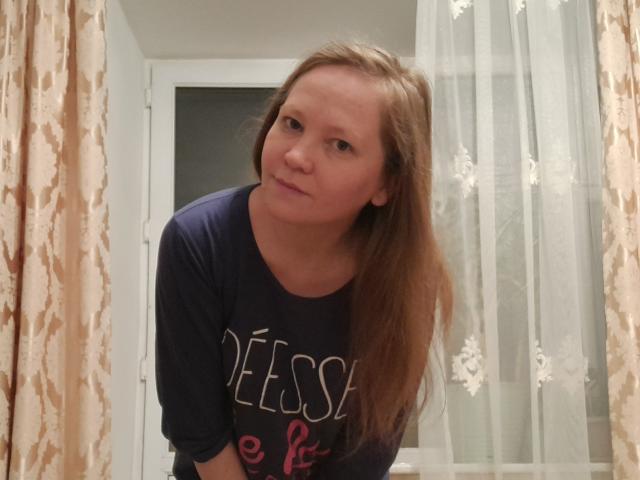LolaAlina with long brown hair, blue t-shirt, leaning forward, slightly smirking, in a room with beige curtains and white lace curtains.
