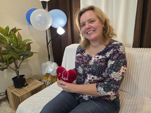 VestaWoody with blonde hair, floral shirt, and jeans, sitting on a white couch, holding a red heart-shaped pillow, smiling, blue and white balloons in the background.