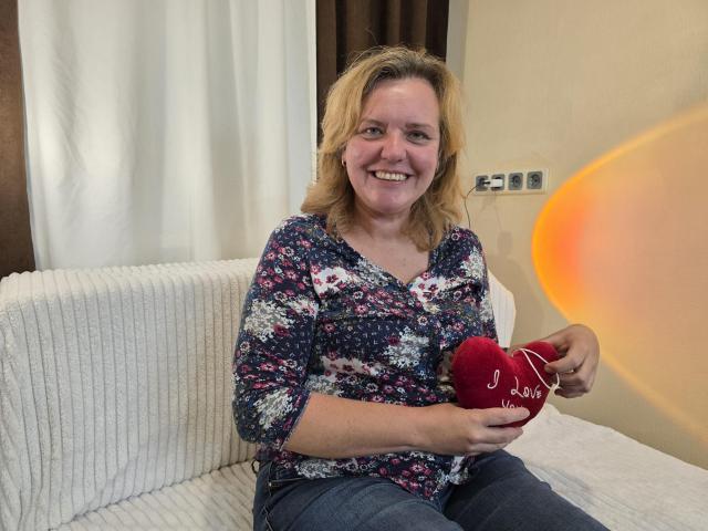 VestaWoody, with blonde hair and a floral shirt, smiles while holding a red heart-shaped pillow with white string. She's sitting on a white ribbed couch.