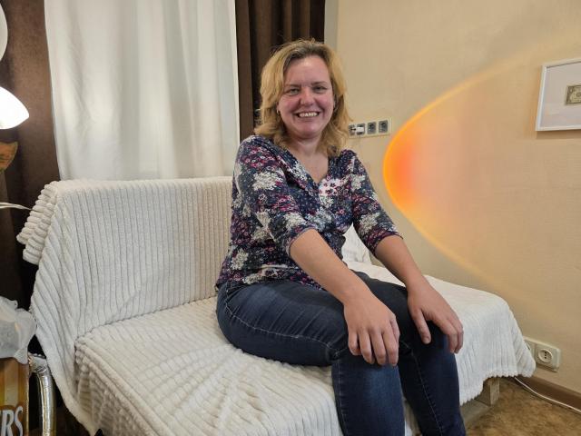 VestaWoody with blonde hair, smiling, wearing a floral shirt, blue jeans, sitting on a white-covered couch, arms resting on knees, orange light reflection on wall.