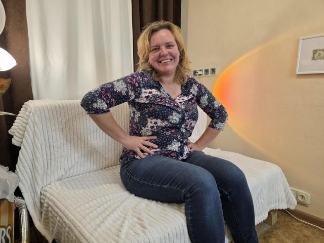 VestaWoody with blonde hair, floral shirt, and blue jeans, sitting on a white-covered couch, smiling, hands on hips, showcasing her medium-sized tits and cute ass.