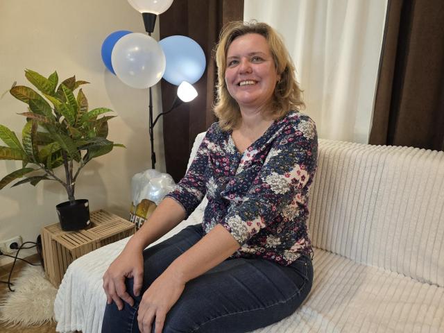 VestaWoody, a middle-aged woman with blonde hair, floral shirt, and dark jeans, sits smiling on a white ribbed couch, with balloons and a plant in the background.